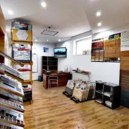 Wnętrze składu budowlanego z ekspozycją płytek elewacyjnych i okładzin ściennych, regały z katalogami i próbkami, biurko z komputerem w tle.