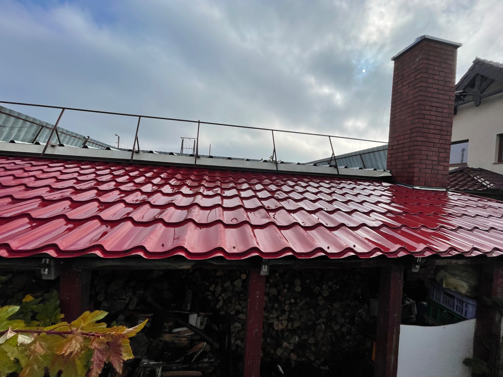 Błyszczący, czerwony dach z blachodachówki trapezowej, widoczny z dołu, z kominem w tle i stosem drewna pod zadaszeniem.