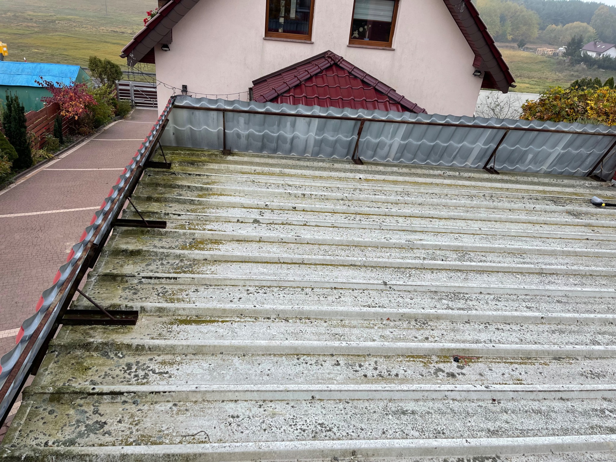 Zabrudzony, pofałdowany dach z metalu z widocznymi plamami mchu i zabrudzeń, z fragmentem domu w tle i widokiem na okolicę w pochmurny dzień.