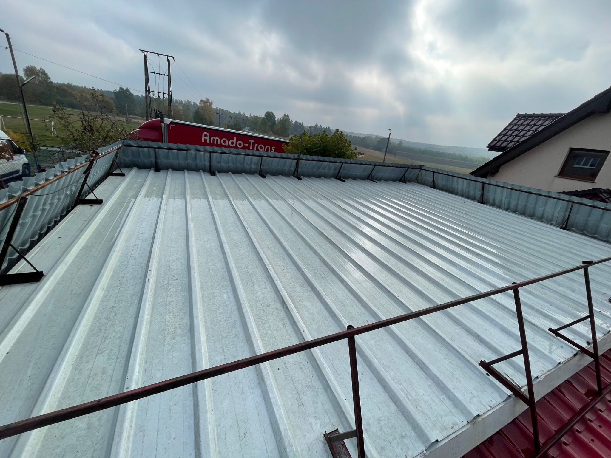 Metalowy dach po umyciu, widoczne mokre fragmenty i zabezpieczenia przed upadkiem, w tle budynek i ciężarówka Amado-Trans.