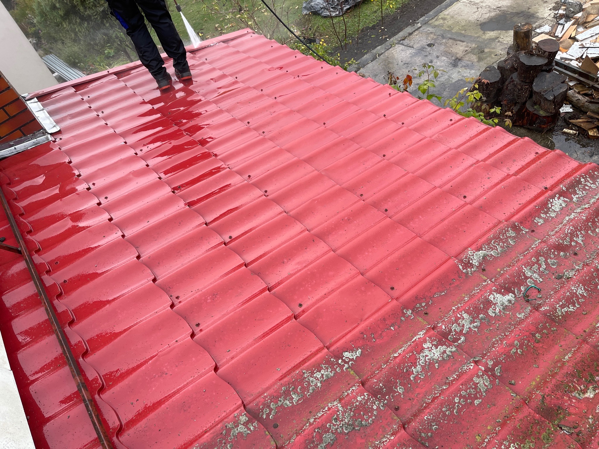 Czerwona dachówka ceramiczna częściowo umyta, z widocznymi śladami zabrudzeń i mchu przed czyszczeniem, oraz pracownik z lancą ciśnieniową w trakcie pracy.