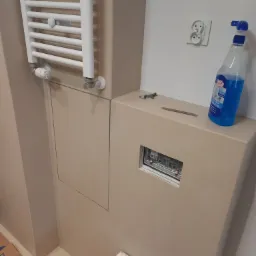 Fragment łazienki w trakcie prac wykończeniowych: beżowe płytki, biały grzejnik drabinkowy, gniazdko elektryczne, otwór w zabudowie z widoczną instalacją, butelka płynu do mycia szyb.