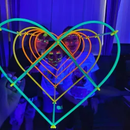 Dzieci z fluorescencyjnym makijażem twarzy pozują za sercem zrobionym ze świecących pałeczek w ciemnym pomieszczeniu z niebieskim oświetleniem.