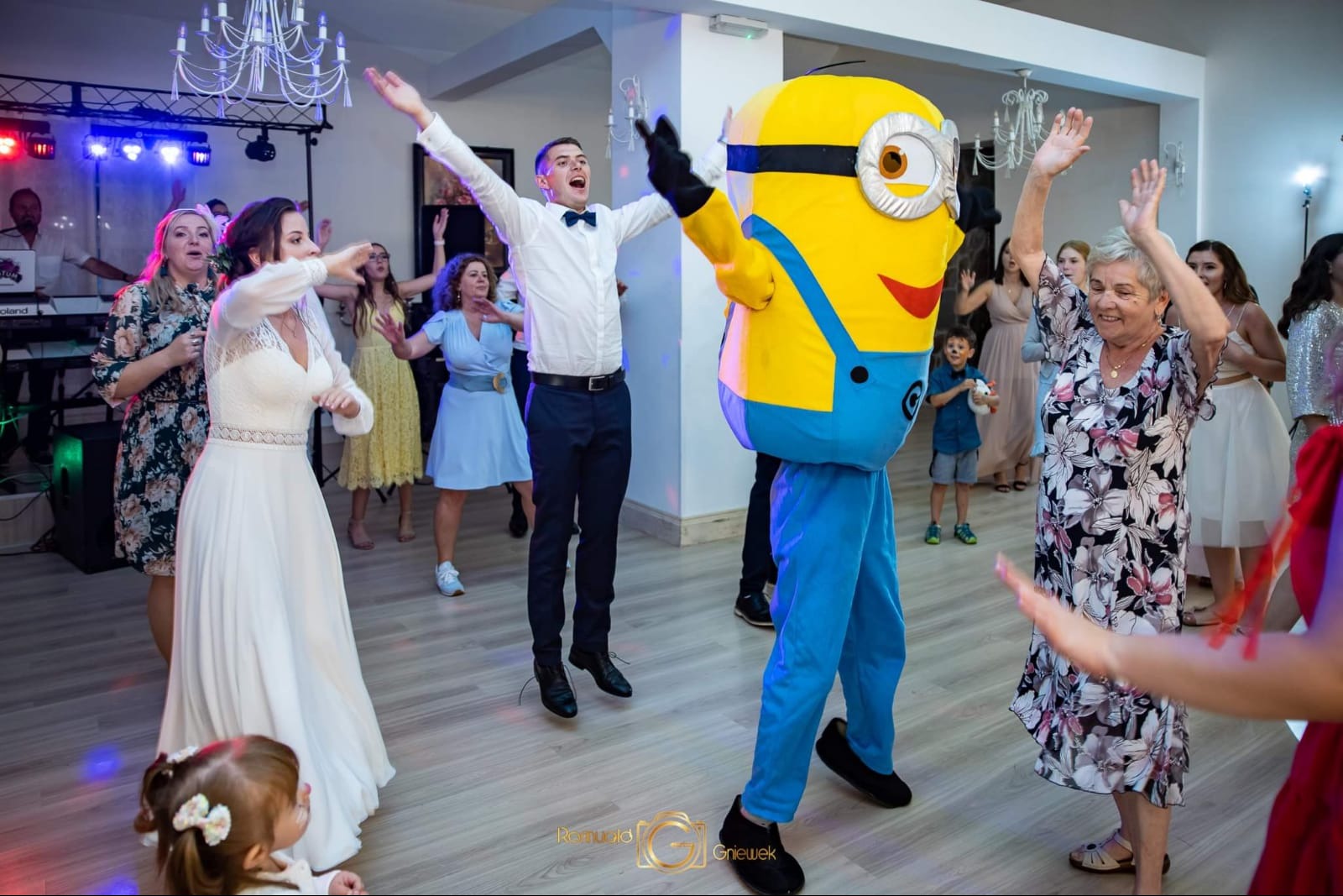 Wnętrze sali bankietowej podczas wesela: panna młoda w białej sukni, goście, mężczyzna w kostiumie Minionka i skaczący konferansjer w muszce.