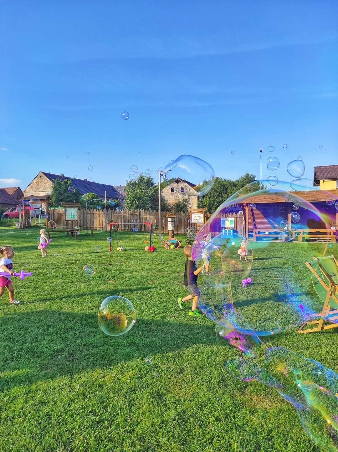 Dzieci na trawniku puszczają duże bańki mydlane, widoczne drewniane budynki w tle, błękitne niebo.