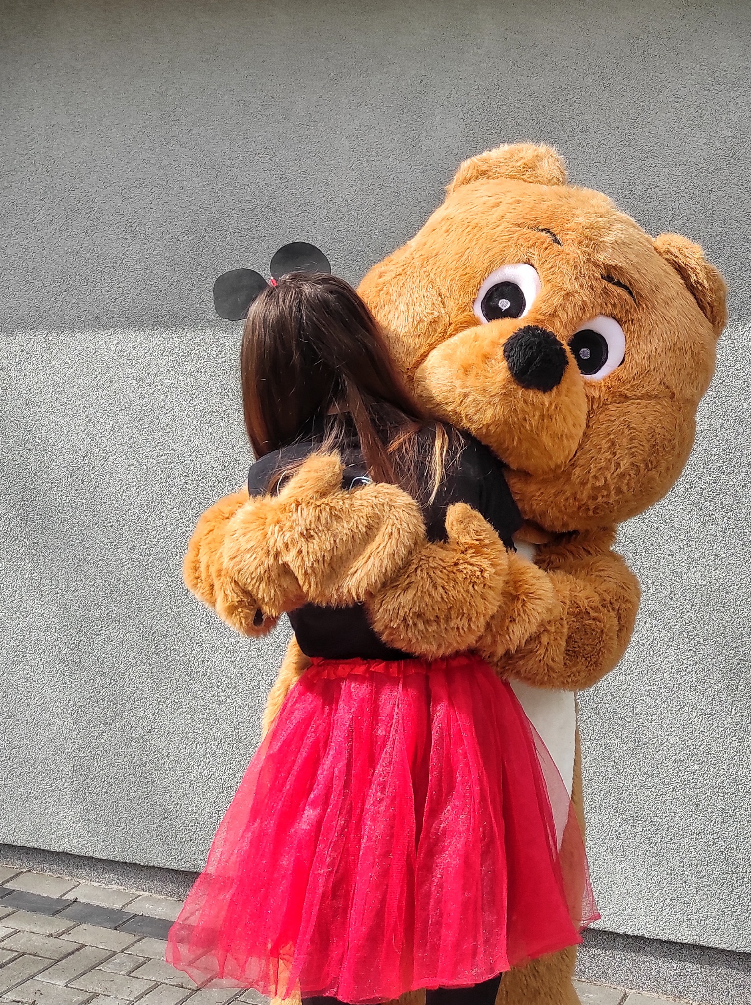 Dziewczynka w czerwonej tiulowej spódnicy przytula się do pluszowego misia-animatora na tle szarej ściany.