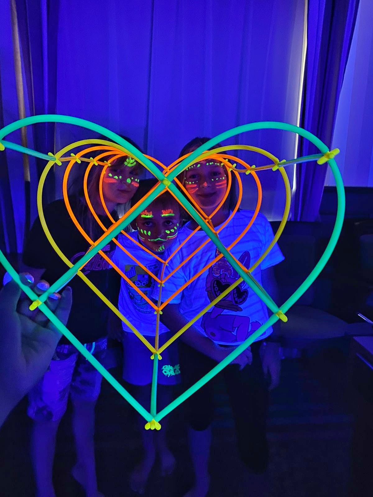 Dzieci z fluorescencyjnym makijażem twarzy pozują za sercem zrobionym ze świecących pałeczek w ciemnym pomieszczeniu z niebieskim oświetleniem.