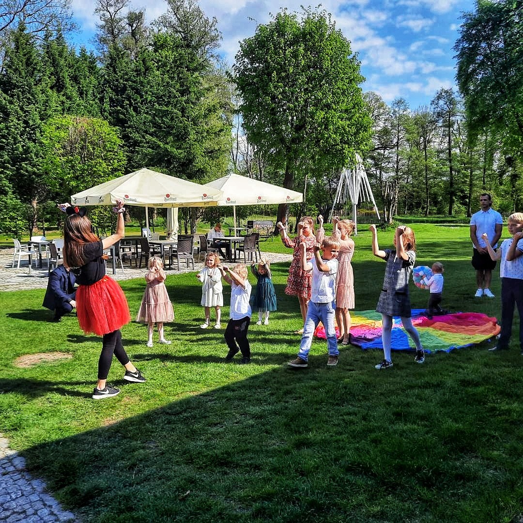 Grupa dzieci bawiących się na trawie podczas imprezy plenerowej, prowadzona przez animatorkę w czerwonej spódnicy tiulowej i opasce z uszami. W tle białe parasole i drzewa.