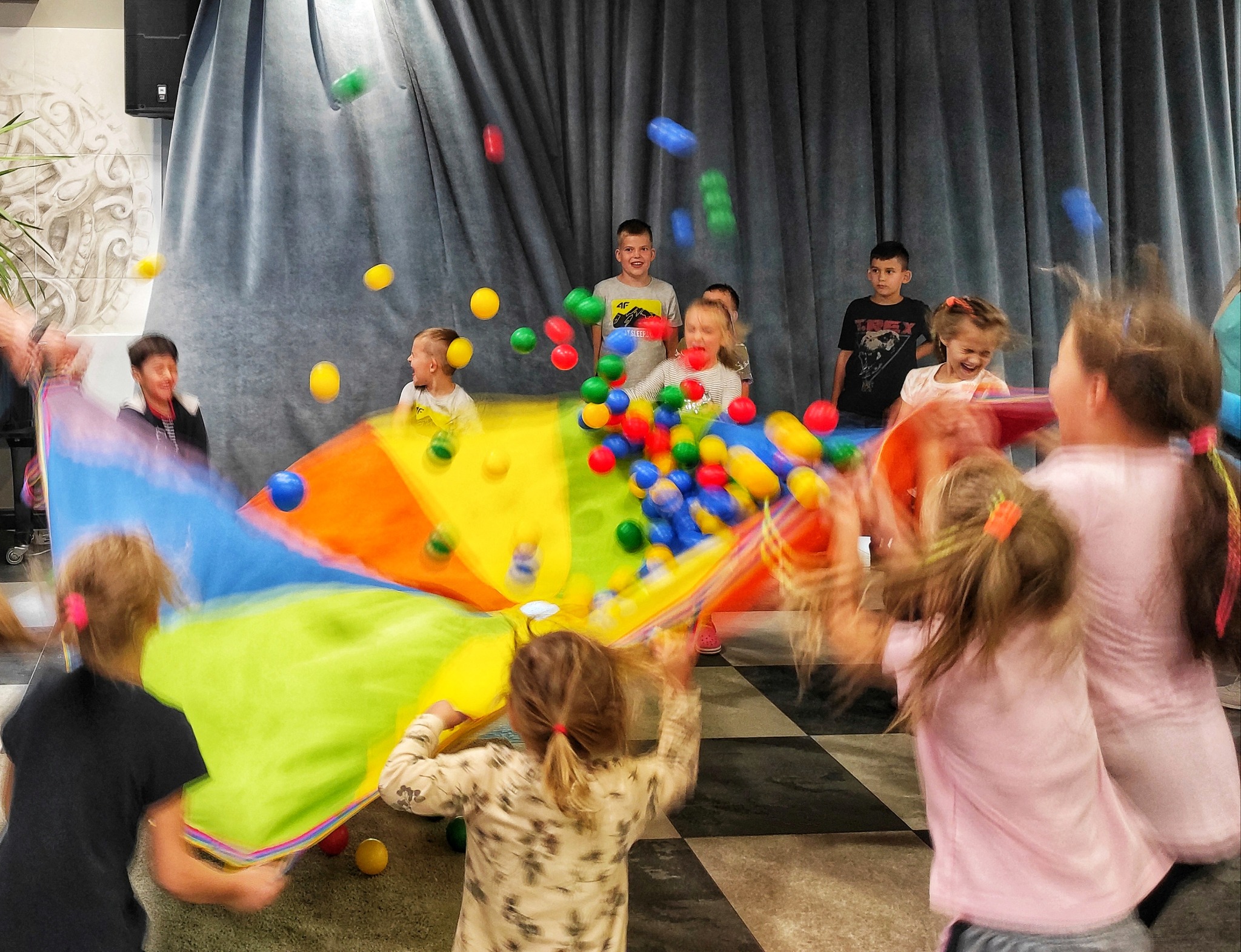 Grupa dzieci bawi się kolorową chustą animacyjną, podrzucając w górę kolorowe piłeczki w sali z szaro-niebieskimi zasłonami w tle.