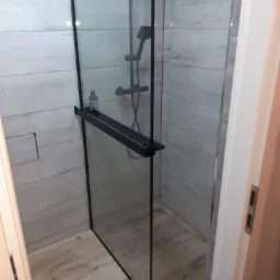 Nowoczesna kabina prysznicowa z czarnymi profilami, jasne płytki imitujące drewno na ścianach i podłodze, półka z kosmetykiem.