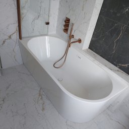 Tomasz Słowik - Nowoczesna, biała wanna z miedzianą armaturą na tle marmurowych płytek w odcieniach bieli i czerni, widoczna kabina prysznicowa z miedzianymi ramami.