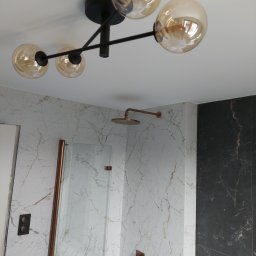 Tomasz Słowik - Nowoczesna łazienka z marmurowymi płytkami w odcieniach bieli i czerni, deszczownicą i baterią w kolorze miedzi oraz designerską lampą sufitową z czterema szklanymi kulami.