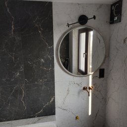 Tomasz Słowik - Wykończona łazienka z czarno-białymi marmurowymi płytkami, okrągłym lustrem z oświetleniem i złotą baterią umywalkową; widoczna niezamontowana kratka wentylacyjna.