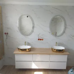 Nowoczesna łazienka z marmurowymi płytkami, owalnymi lustrami z podświetleniem LED, złotymi umywalkami i grzejnikiem drabinkowym w kolorze złota.