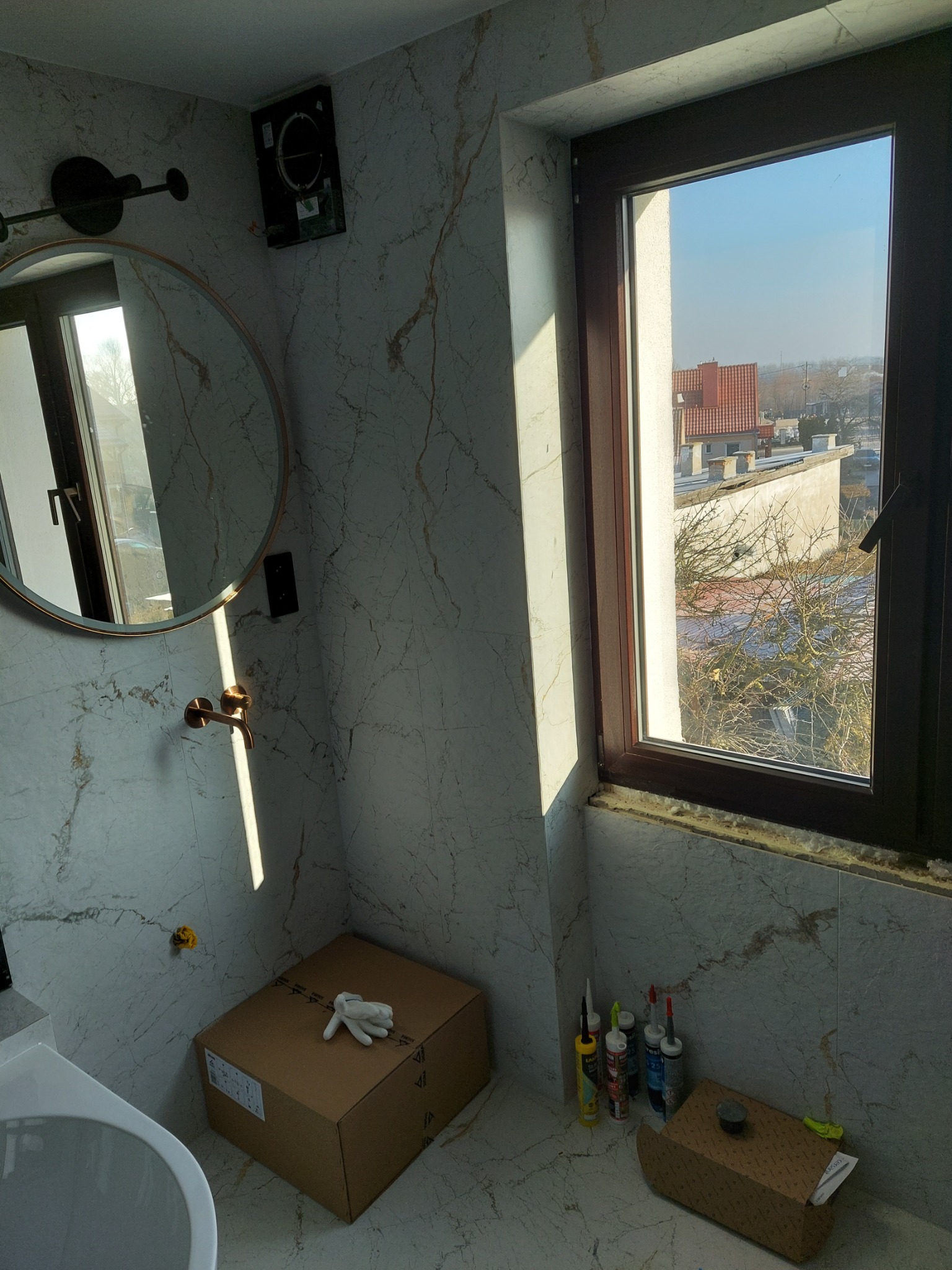 Fragment łazienki w trakcie wykańczania: marmurowe płytki, okrągłe lustro, złota armatura, karton z rękawicami roboczymi, tuby silikonu przy oknie z widokiem na zabudowę miejską.