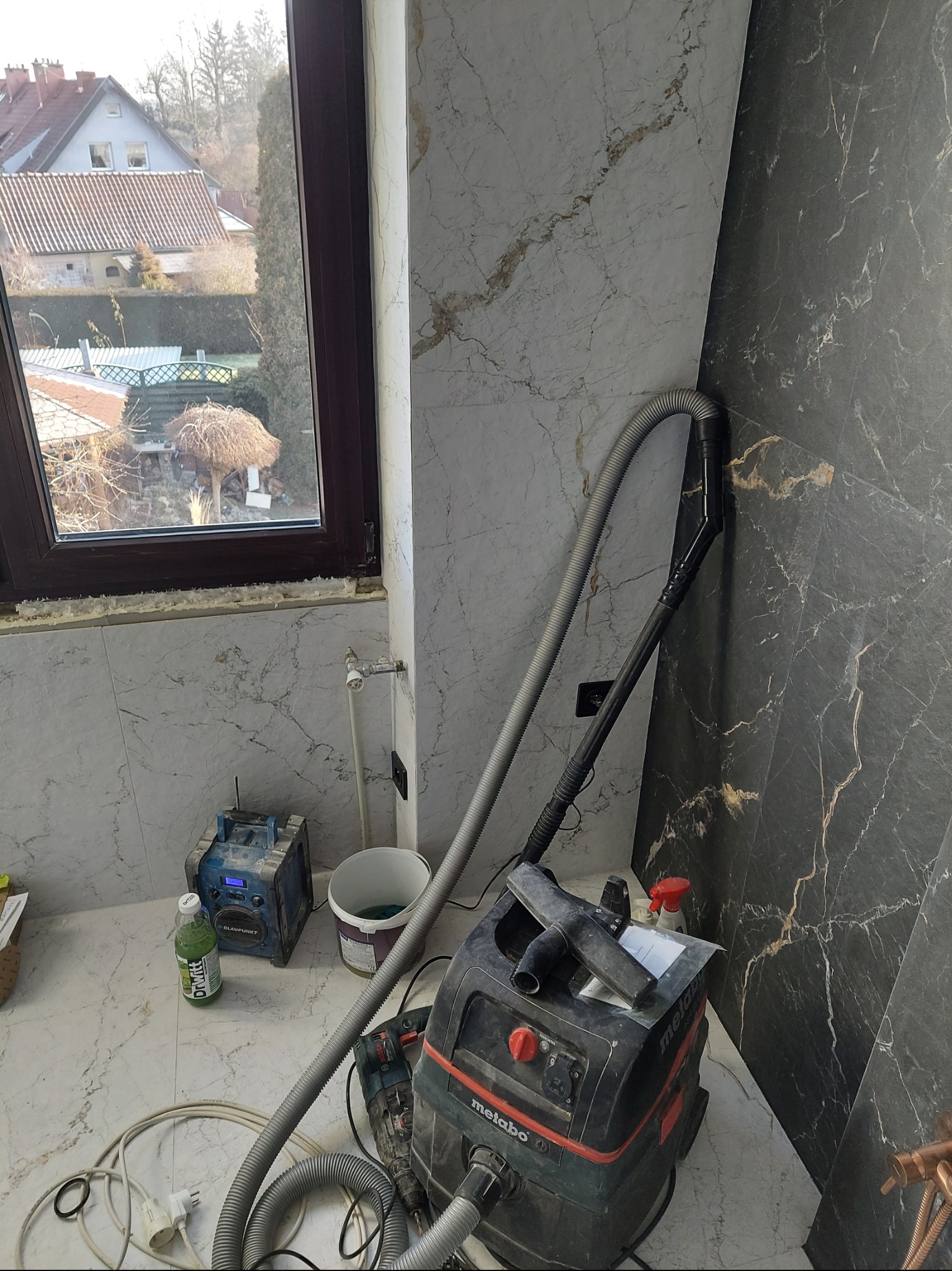 Łazienka w trakcie remontu: widoczny odkurzacz przemysłowy Metabo, radio budowlane Blaupunkt, biało-czarne marmurowe płytki ścienne, okno z widokiem na ogród i domy.