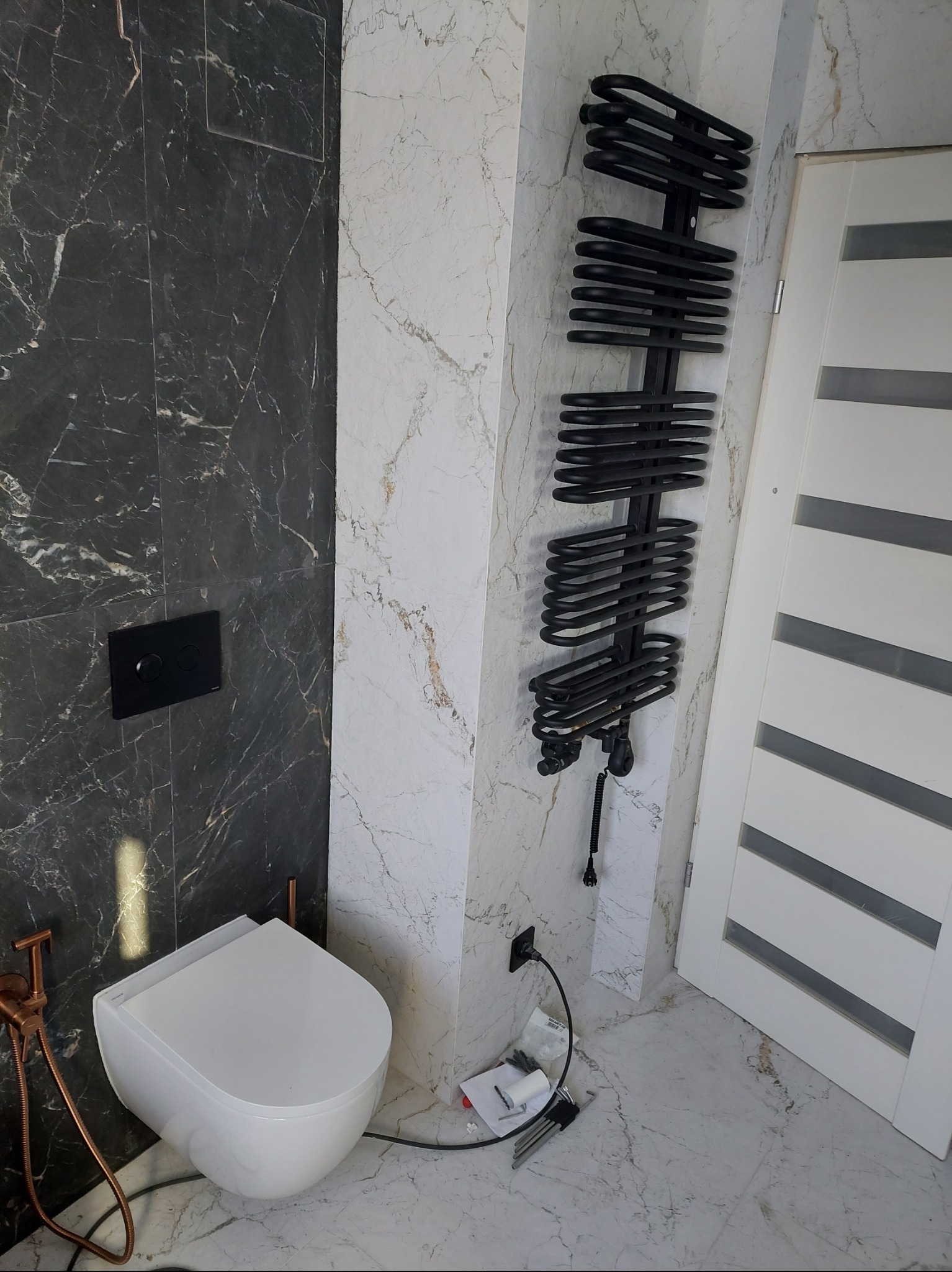Nowoczesna łazienka w trakcie wykańczania: podwieszana toaleta z miedzianą bidetką, czarny grzejnik drabinkowy i marmurowe płytki na ścianach, widoczne narzędzia i kable na podłodze.