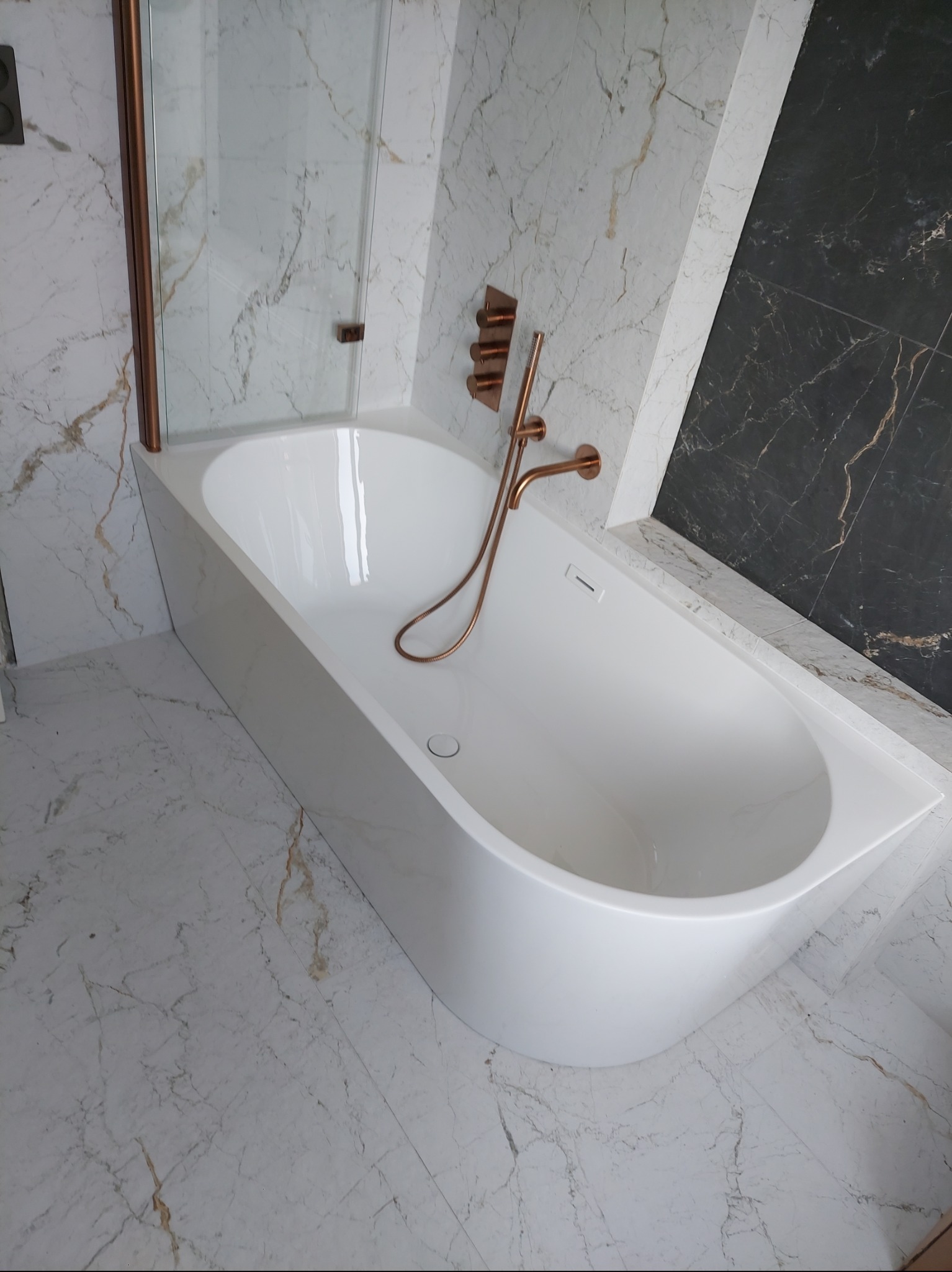 Nowoczesna, biała wanna z miedzianą armaturą na tle marmurowych płytek w odcieniach bieli i czerni, widoczna kabina prysznicowa z miedzianymi ramami.