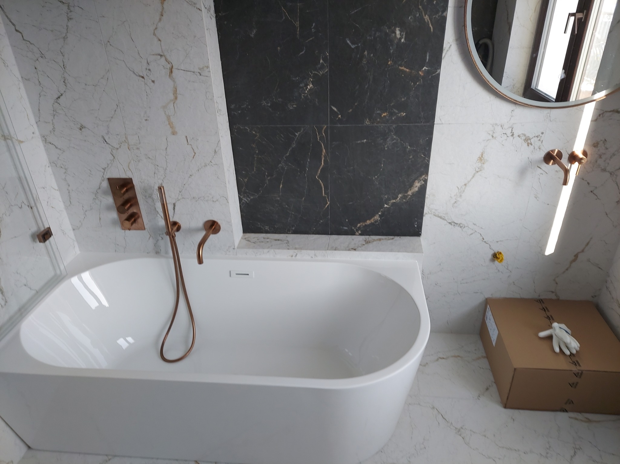 Nowoczesna łazienka z białą wanną, marmurowymi płytkami w odcieniach bieli i czerni, armaturą w kolorze miedzi oraz kartonem z rękawicami na podłodze.