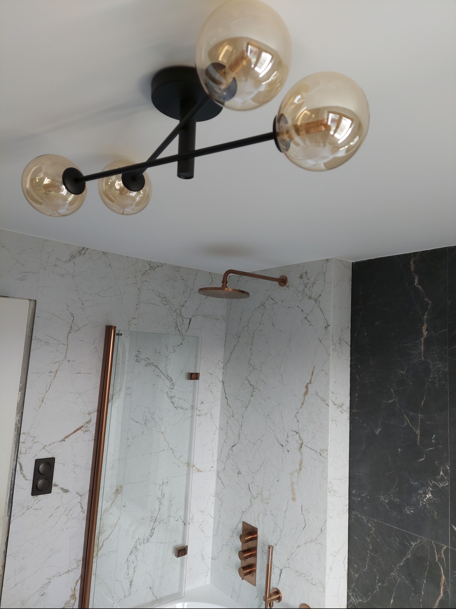 Nowoczesna łazienka z marmurowymi płytkami w odcieniach bieli i czerni, deszczownicą i baterią w kolorze miedzi oraz designerską lampą sufitową z czterema szklanymi kulami.