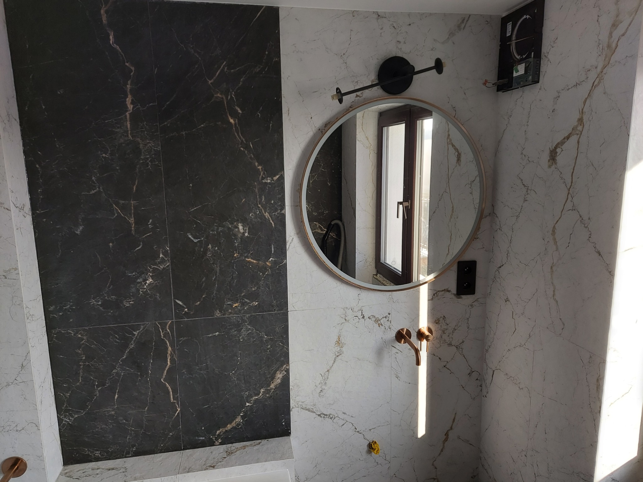 Wykończona łazienka z czarno-białymi marmurowymi płytkami, okrągłym lustrem z oświetleniem i złotą baterią umywalkową; widoczna niezamontowana kratka wentylacyjna.