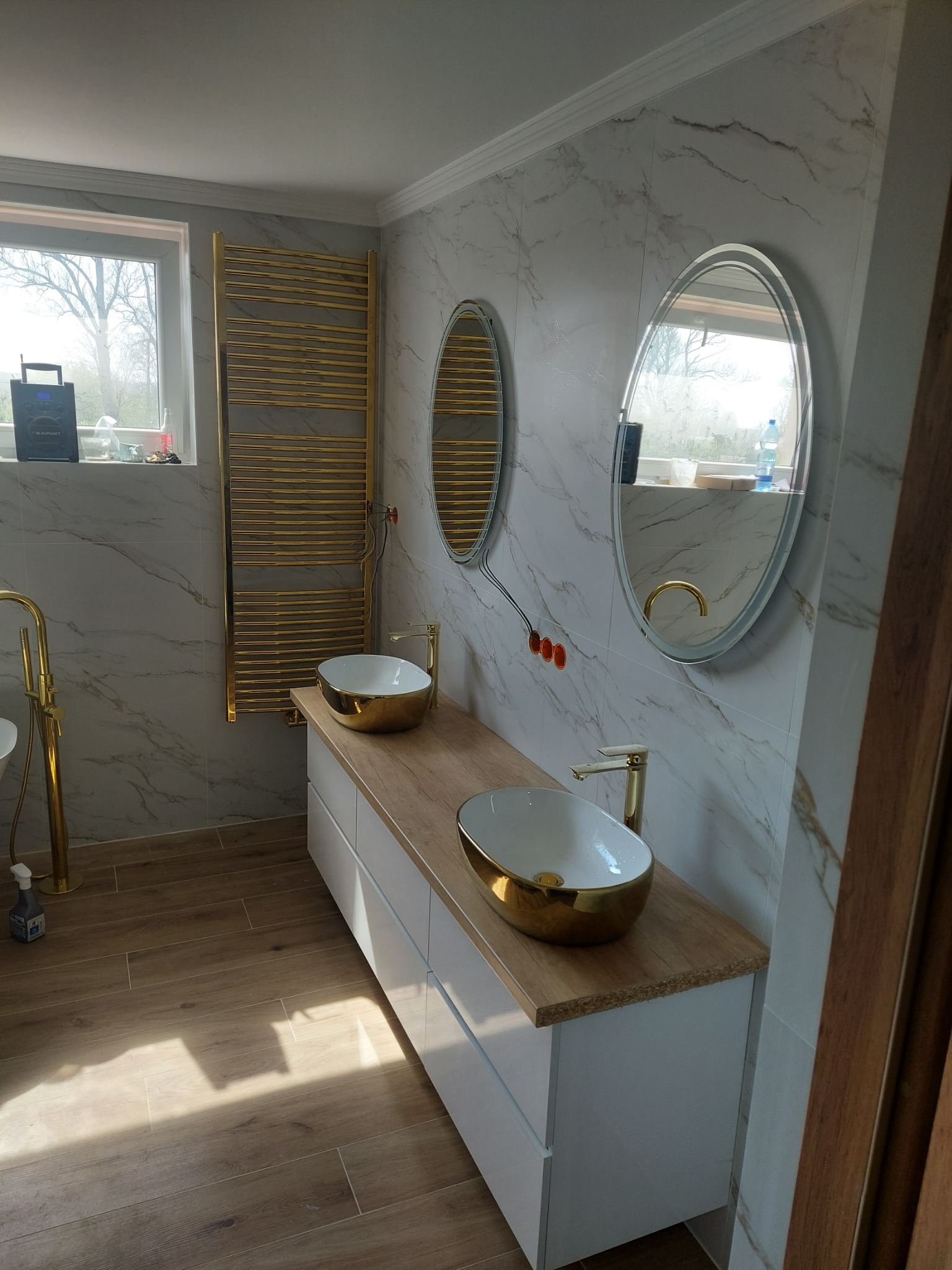 Elegancka łazienka: marmurowe ściany, drewnopodobna podłoga, biała szafka z drewnianym blatem i dwie złote umywalki. Grzejnik drabinkowy w kolorze złota.