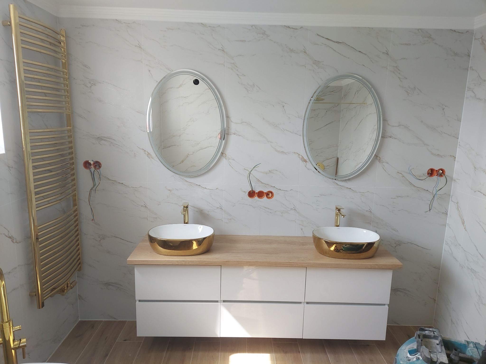 Nowoczesna łazienka z marmurowymi płytkami, owalnymi lustrami z podświetleniem LED, złotymi umywalkami i grzejnikiem drabinkowym w kolorze złota.