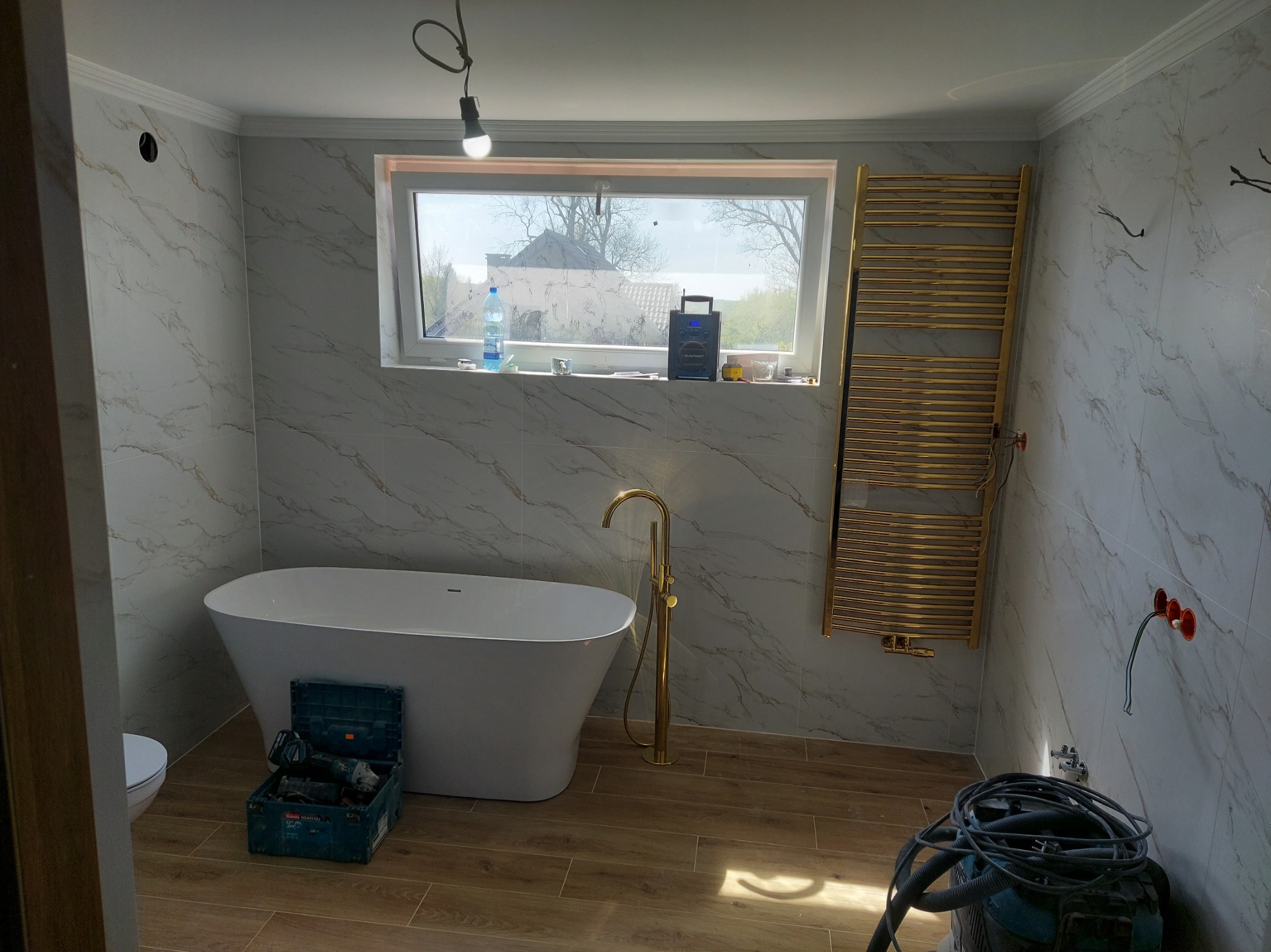 Biała wanna wolnostojąca i złota armatura w łazience z marmurowymi płytkami. Na podłodze panele imitujące drewno. Widoczny grzejnik drabinkowy i narzędzia.