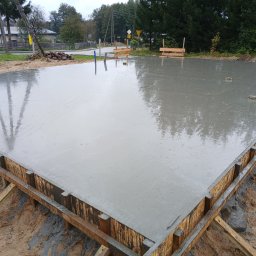 KARKAM AGRAR SERVICE KAMIL LEWICKI - Zalany betonem szalunek fundamentu domu jednorodzinnego, widoczne zbrojenia i kotwy, w tle drzewa i zabudowania.