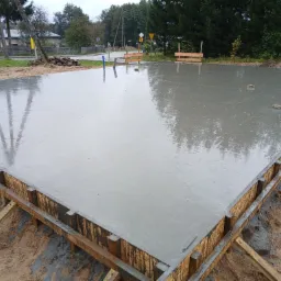 Zalany betonem szalunek fundamentu domu jednorodzinnego, widoczne zbrojenia i kotwy, w tle drzewa i zabudowania.