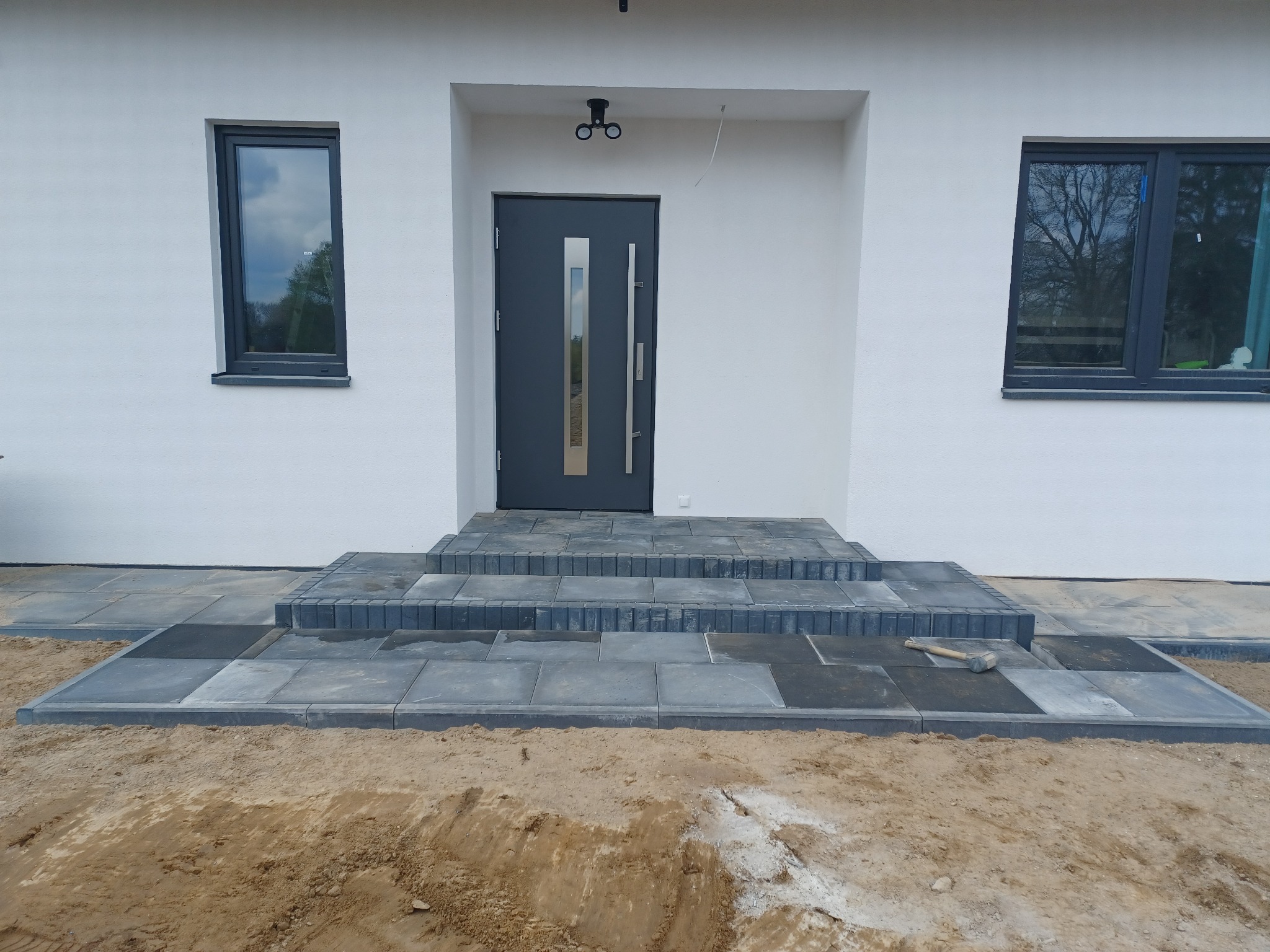 Wejście do nowoczesnego domu z szarymi płytami brukowymi tworzącymi schody i chodnik; widoczny młotek brukarski na jednej z płyt, sugerujący trwające prace wykończeniowe.