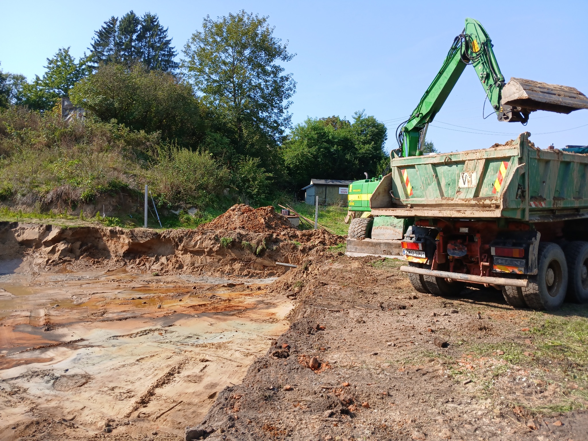 Zielona koparka ładuje ziemię do wywrotki na tle wykopu z widocznymi warstwami gleby i drzewami w tle.