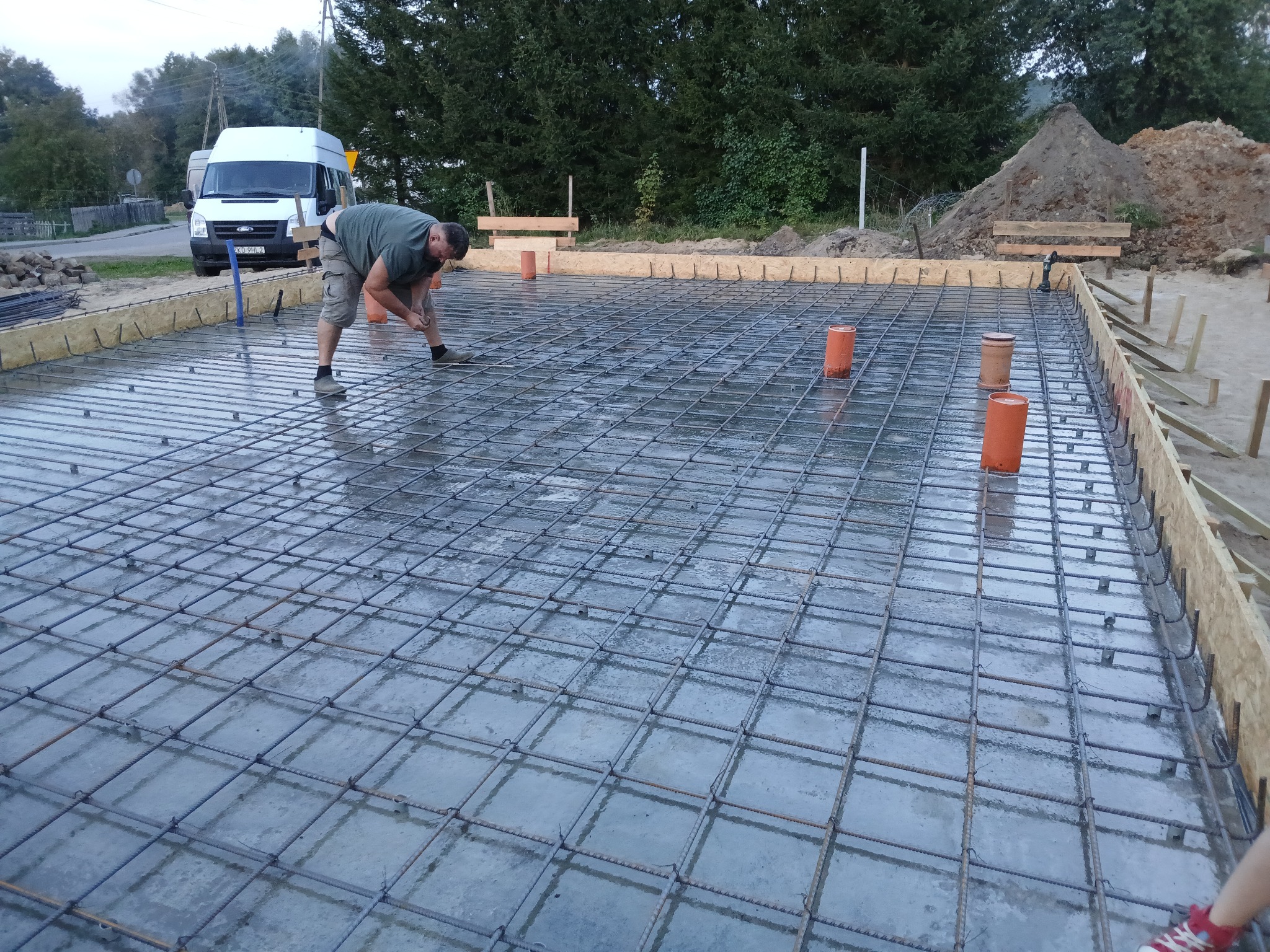 Zbrojenie płyty fundamentowej z wystającymi rurami kanalizacyjnymi, widoczny pracownik w trakcie montażu, szalunek z desek, w tle biały bus i hałda ziemi.