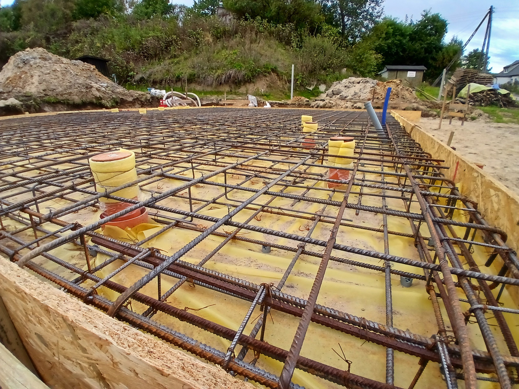 Zbrojenie fundamentu domu jednorodzinnego, widoczne pręty stalowe połączone drutem, żółta folia izolacyjna, szalunek drewniany, w tle hałda ziemi i zieleń.