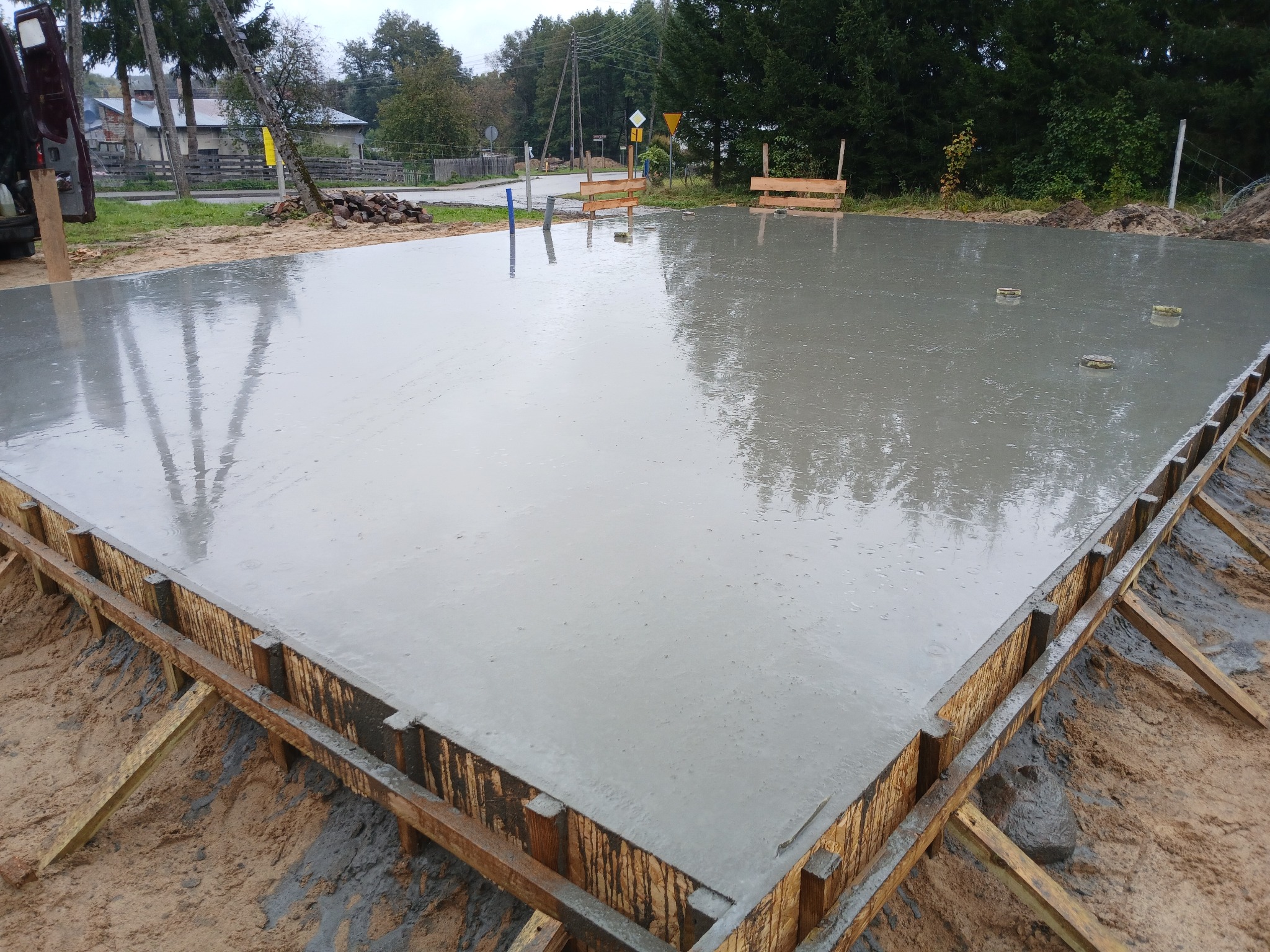 Zalany betonem szalunek fundamentu domu jednorodzinnego, widoczne zbrojenia i kotwy, w tle drzewa i zabudowania.