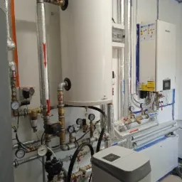 Kotłownia z białym zasobnikiem ciepłej wody, licznymi rurami, manometrami i urządzeniem do uzdatniania wody, widoczne przewody w izolacji termicznej i biały piec grzewczy na ścianie.