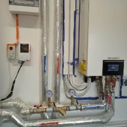 Węzeł cieplny z kotłem grzewczym, rurami w izolacji aluminiowej oznaczonymi strzałkami kierunku przepływu, manometrami, zaworami i elektrycznym panelem sterowania na białej ścianie.