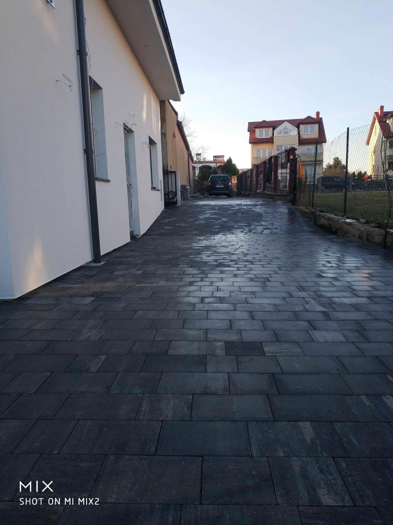 Długa, wybrukowana podjazd z ciemnej kostki prowadząca do domu, widok z perspektywy chodnika, po lewej stronie biały budynek z orynnowaniem, w tle domy jednorodzinne i samochody.