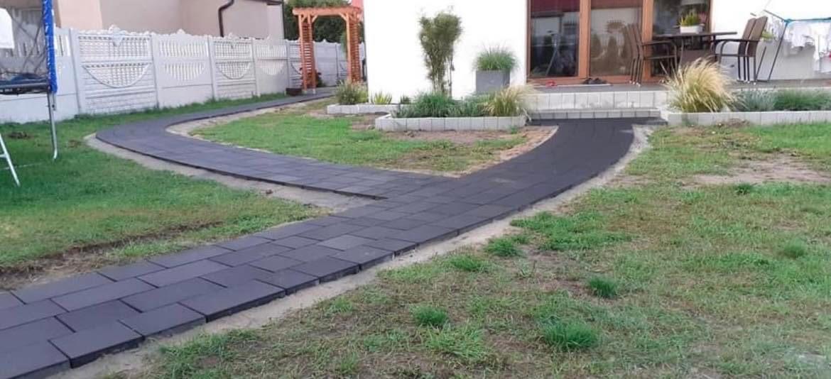 Ścieżka z ciemnej kostki brukowej prowadząca przez trawnik do domu, z widocznym białym ogrodzeniem i drewnianą pergolą w tle.