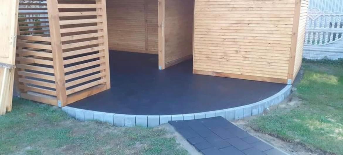 Nowoczesna, drewniana altana z czarną, gładką posadzką obramowaną szarymi kostkami, prowadzącymi do ścieżki z ciemnych płyt.