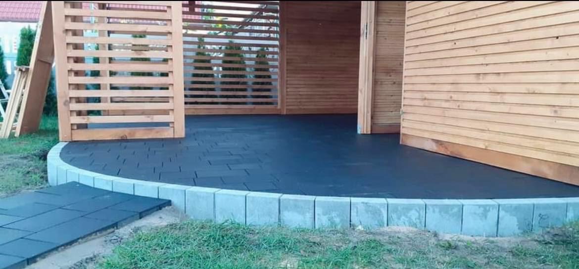 Wykończona, zacieniona powierzchnia z czarnej kostki brukowej w Mrzezinie, otoczona niskim, betonowym obramowaniem, przylegająca do drewnianej konstrukcji.