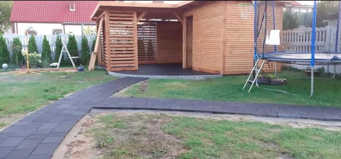 Nowoczesna altana z drewna zadaszona czarną, gumową kostką, do której prowadzi ścieżka z tej samej kostki, obok trampolina.