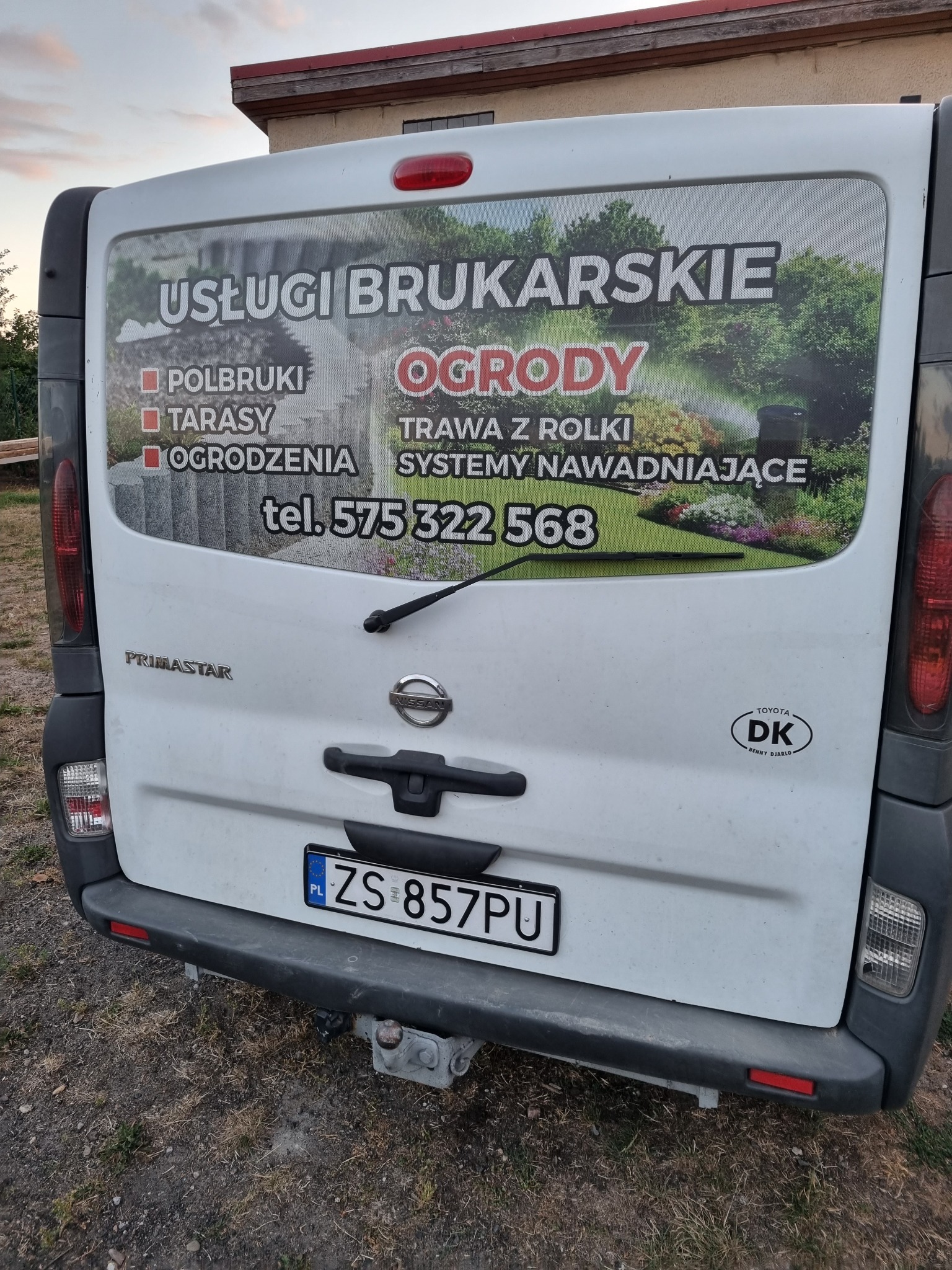 Tył brudnego, białego samochodu dostawczego z reklamą usług brukarskich i ogrodniczych: polbruki, tarasy, ogrodzenia, trawa z rolki, systemy nawadniające. Widoczny numer telefonu kontaktowego.