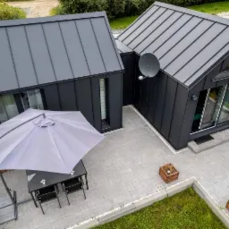 Nowoczesny dom z ciemną elewacją i metalowym dachem, widok z góry na taras z szarą kostką brukową, parasolem i meblami ogrodowymi, oraz satelitą na ścianie.