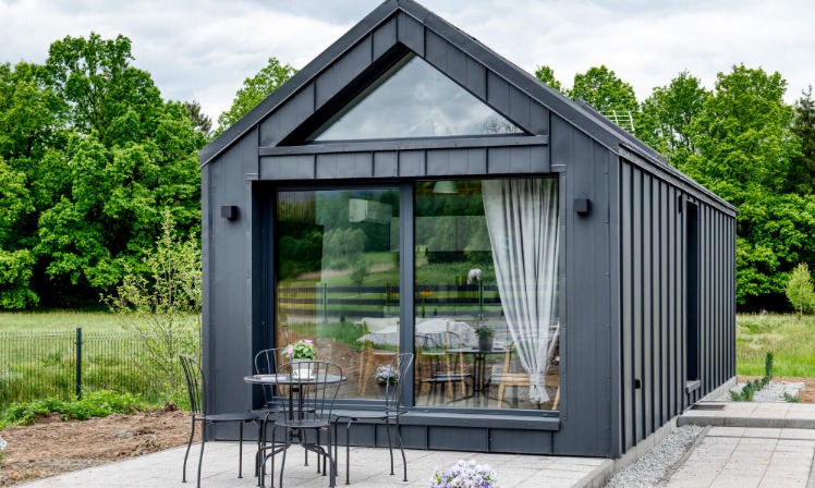 Nowoczesny, grafitowy domek letniskowy z przeszkleniami i trójkątnym oknem na tle zieleni; na pierwszym planie stolik kawowy z krzesłami.
