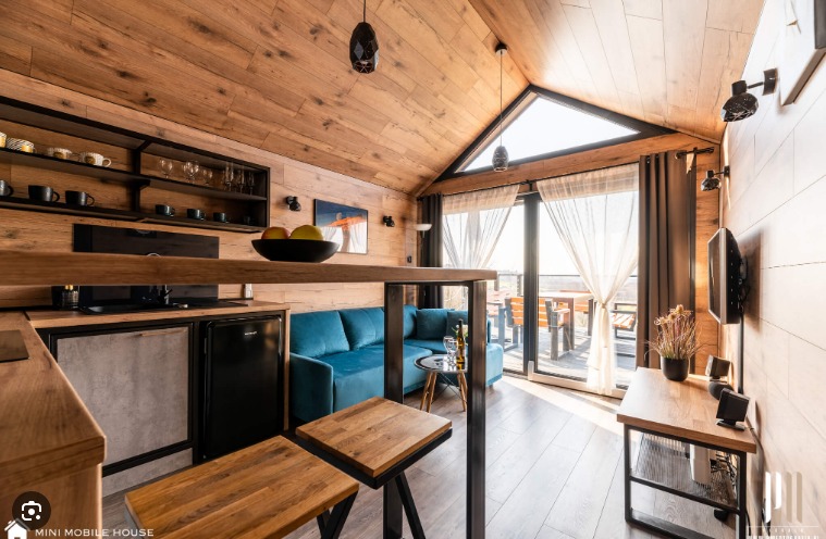 Drewniane wnętrze domku z aneksem kuchennym, turkusową sofą i dużym oknem trójkątnym w szczycie dachu, widok na taras.