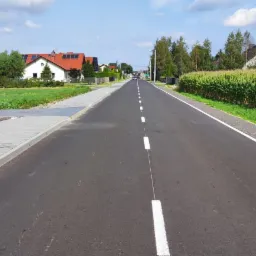 Asfaltowa droga z nowym oznakowaniem poziomym, po lewej stronie chodnik z kostki brukowej, w tle zabudowa jednorodzinna i pole kukurydzy.