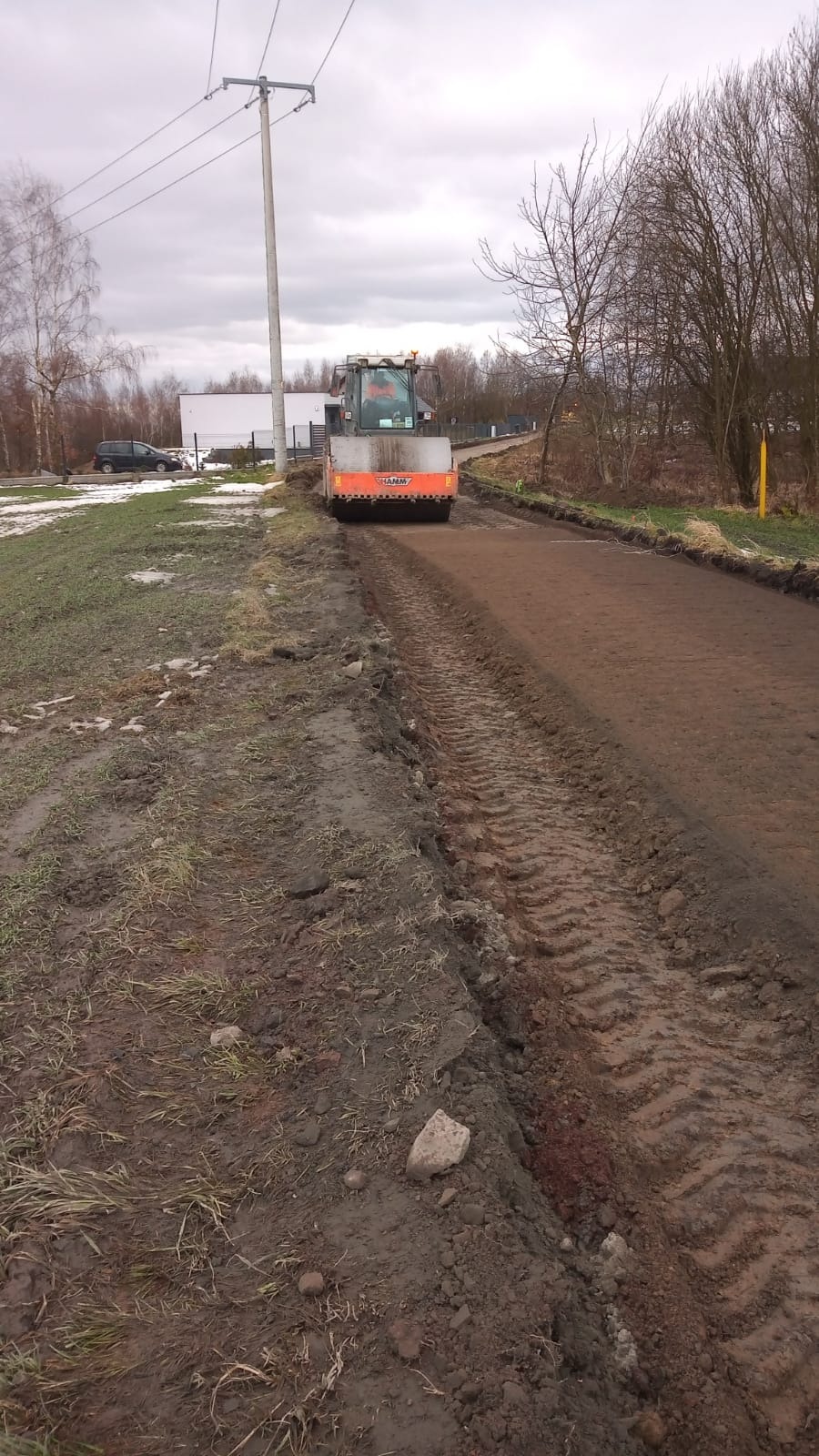 Pomarańczowy walec drogowy HAMM ubija nawierzchnię drogi gruntowej, widoczne ślady opon, obok błotniste pobocze z trawą, słup energetyczny i budynek w tle, pochmurne niebo.