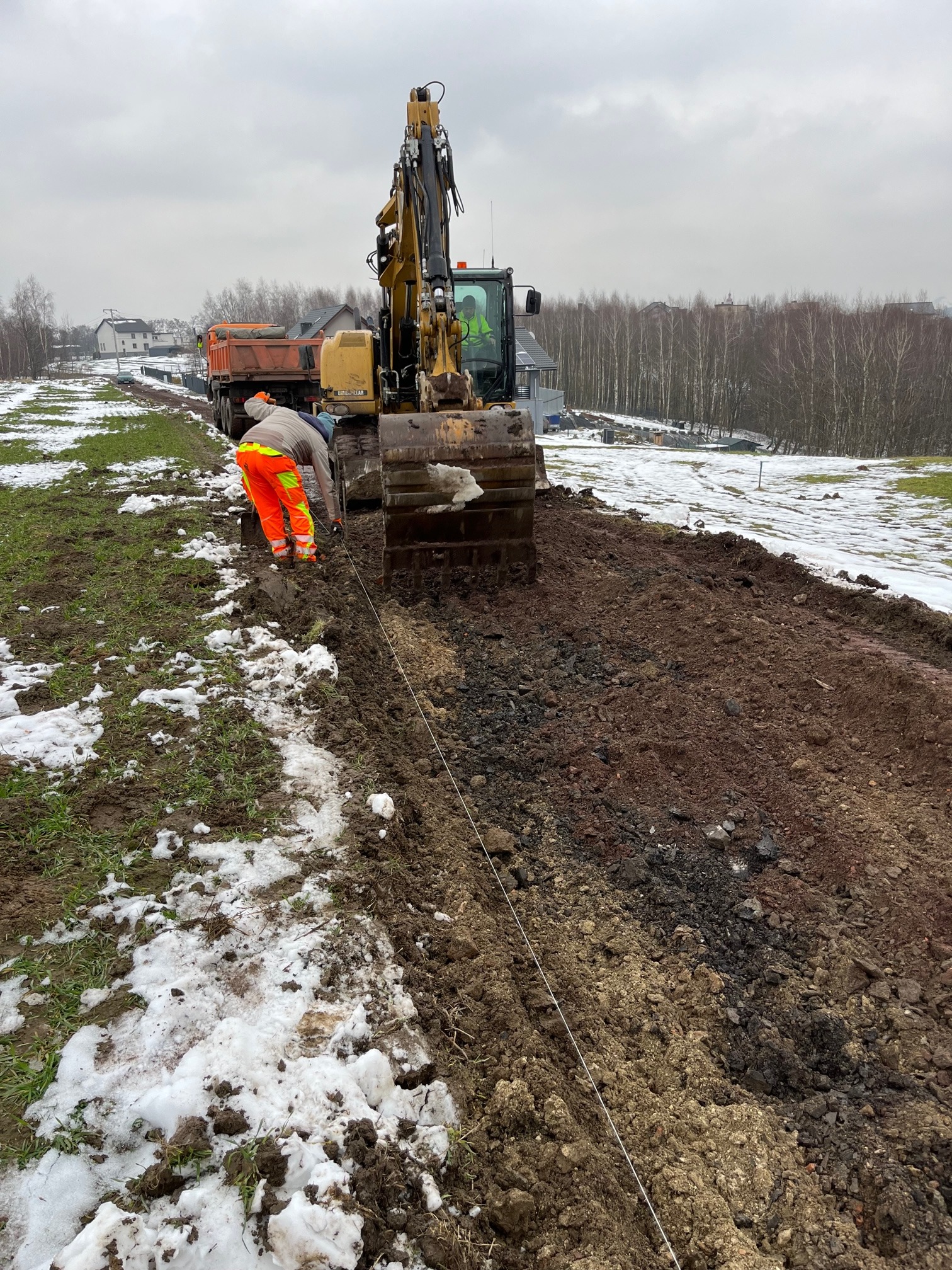 Żółta koparka podczas wykopu rowu w grząskim terenie z resztkami śniegu, robotnik w pomarańczowych spodniach i szarym kombinezonie wyznacza linię, w tle widoczna ciężarówka i zabudowania.