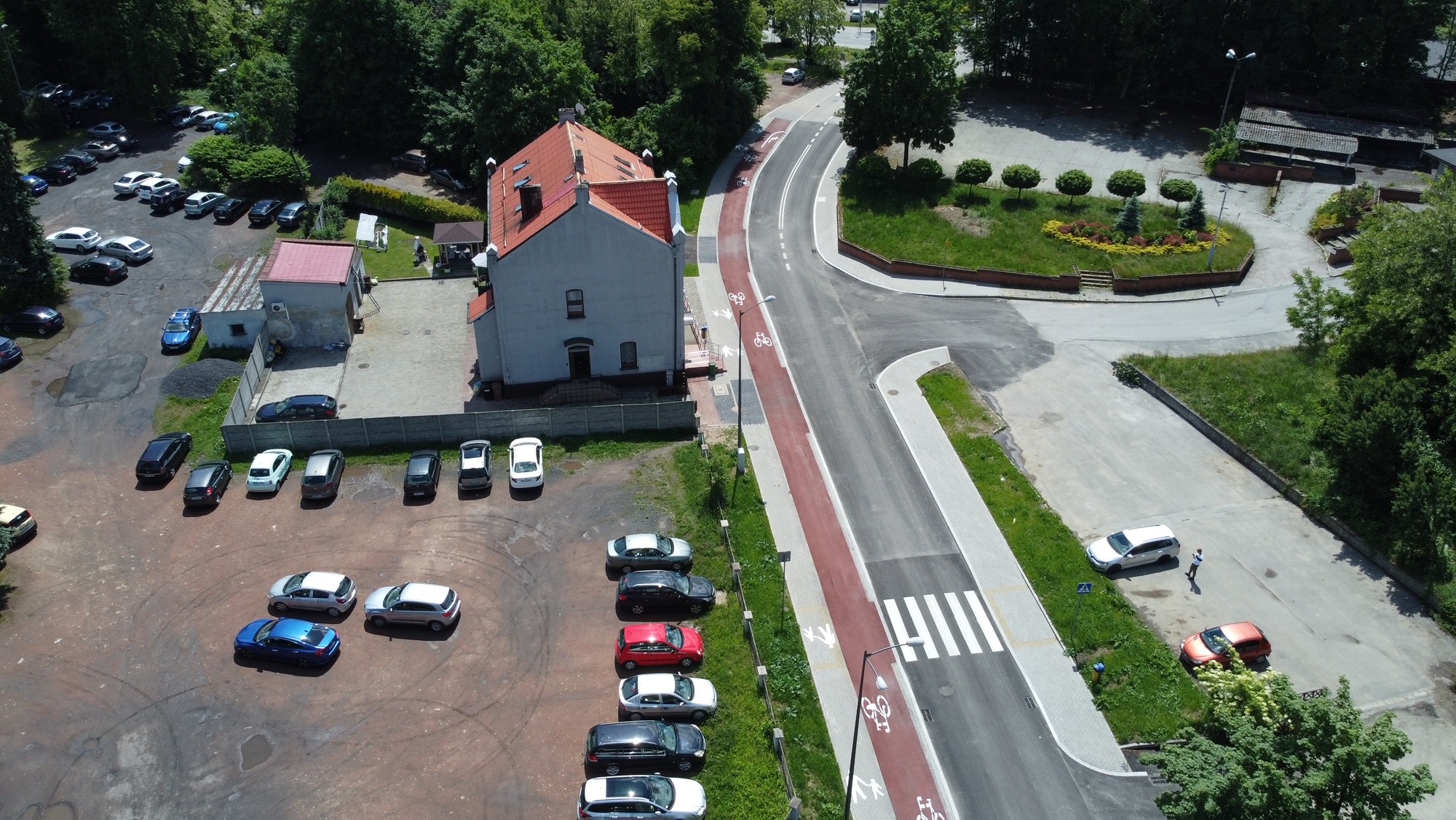 Widok z lotu ptaka na nowo wybudowaną drogę z wydzielonym pasem dla rowerów, parkingiem i budynkiem z czerwonym dachem, otoczone zielenią.